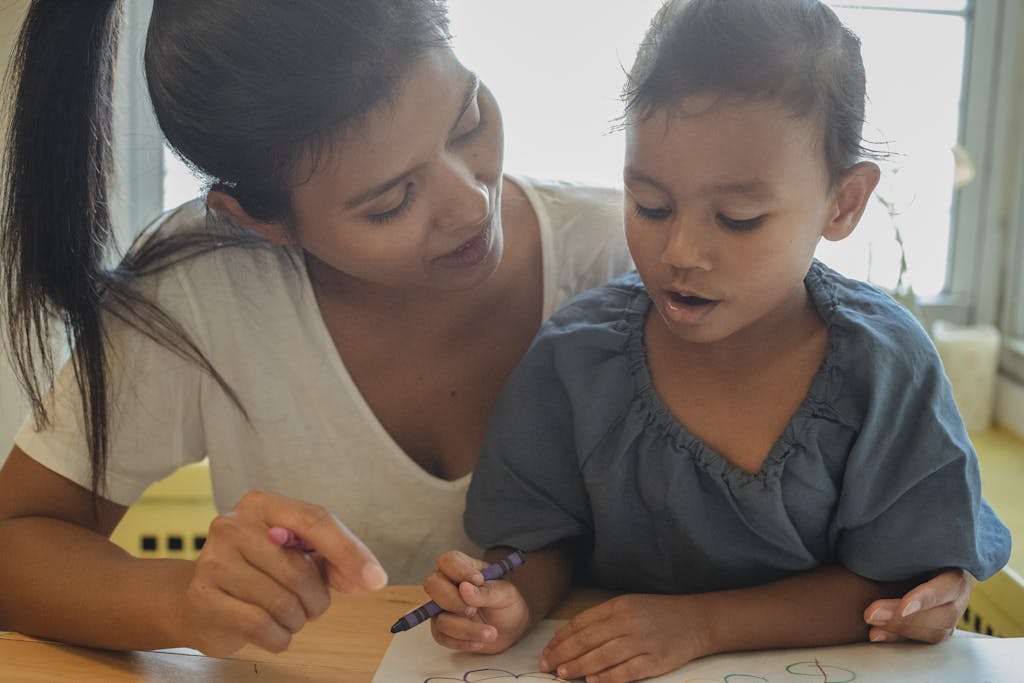 Content young ethnic female mother and cute daughter sitting at table and drawing on paper sheet with crayons while spending time together at home